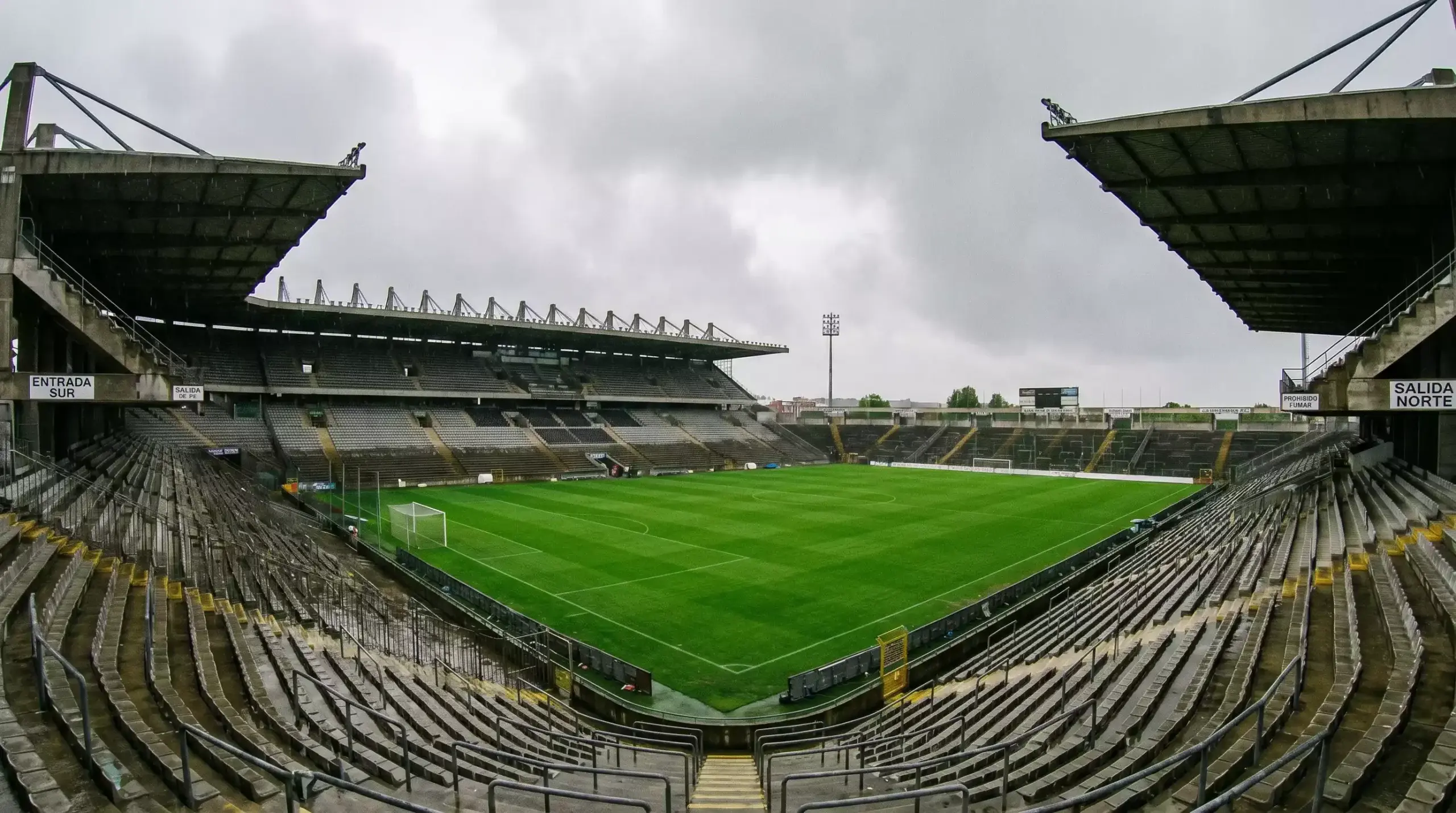 Estadio de fútbol alemán con gradas empinadas y césped verde intenso bajo cielo gris